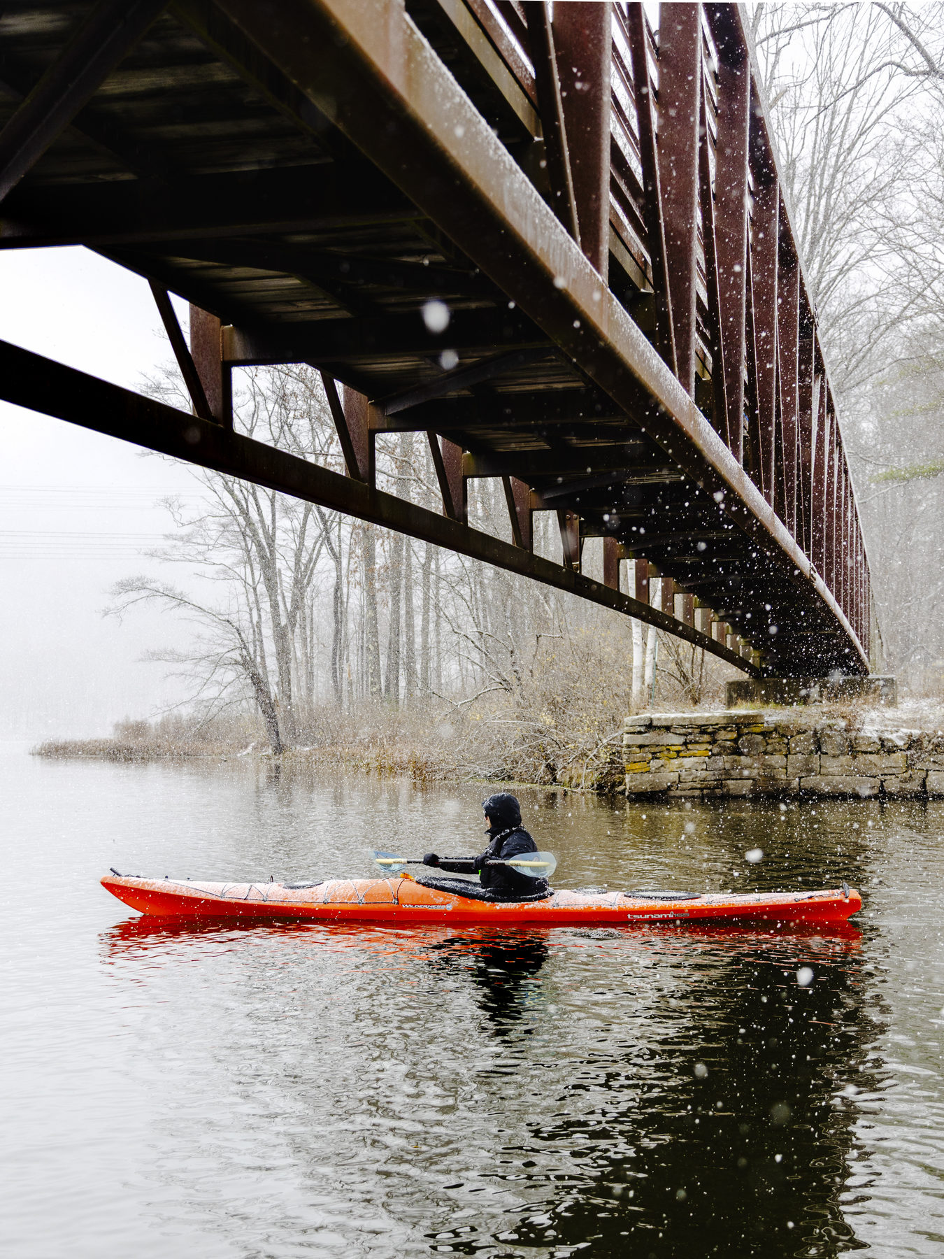 A kayaker paddling under a bridge during a snowfall